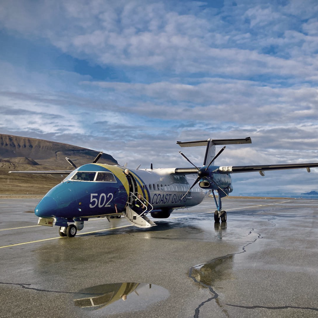 A Coast Guard aircraft labeled '502' parked on a wet runway, with its door open and staircase extended. The backdrop features a mountainous landscape under a cloudy sky, reflecting in the water puddles on the ground.