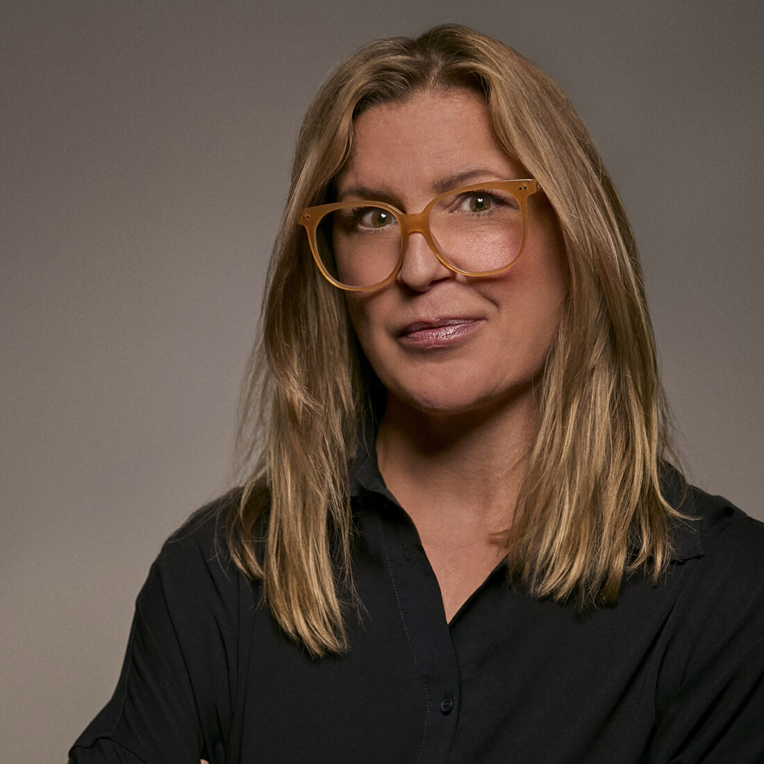 A portrait of a woman with blonde hair wearing oversized glasses and a black shirt, smiling confidently against a neutral background, symbolizing expertise and professionalism in airborne surveillance technology.