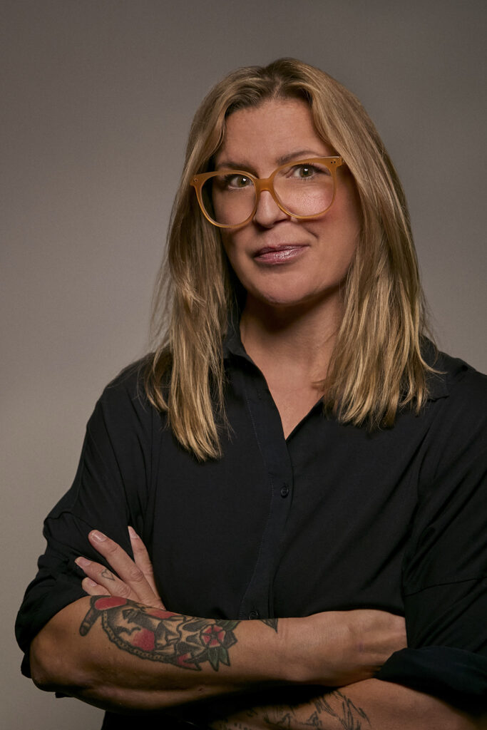 A portrait of a woman with blonde hair wearing oversized glasses and a black shirt, smiling confidently against a neutral background, symbolizing expertise and professionalism in airborne surveillance technology.