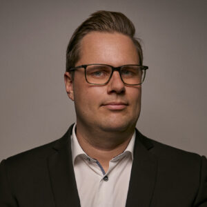 A portrait of a man wearing glasses and a suit, looking confident and professional against a neutral background, representing leadership in airborne surveillance technology.