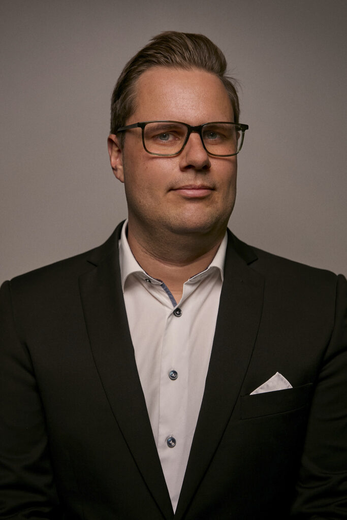 A portrait of a man wearing glasses and a suit, looking confident and professional against a neutral background, representing leadership in airborne surveillance technology.