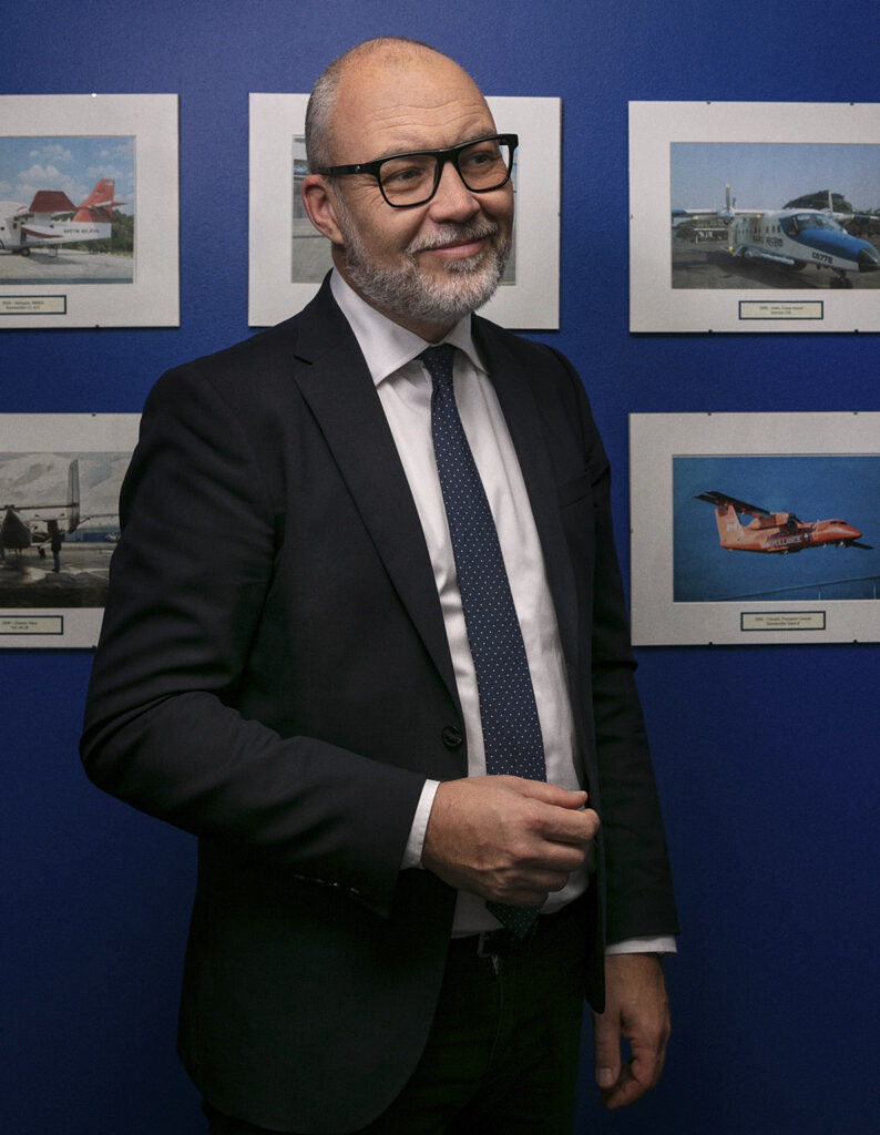 A professionally dressed man standing in front of a blue wall featuring framed images of various aircraft, highlighting expertise and involvement in airborne surveillance technology.